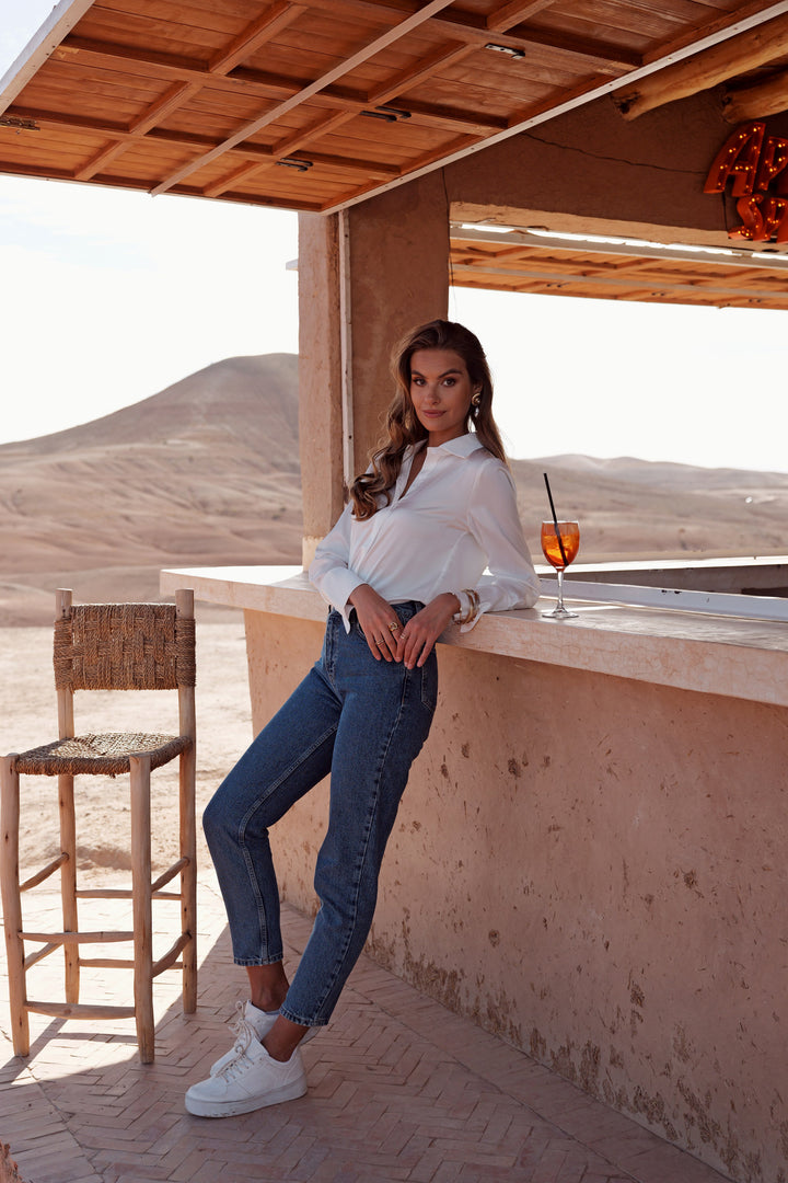 A woman stands on a desert patio under a wooden roof, leaning against a concrete counter. She is wearing a white long-sleeve blouse, blue cropped jeans, and white sneakers.