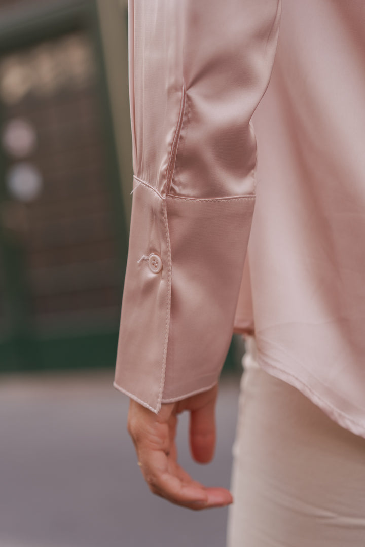 Close-up of a person wearing a light blush pink satin long-sleeve blouse, showing the satin fabric sheen and a buttoned cuff, paired with light beige trousers.