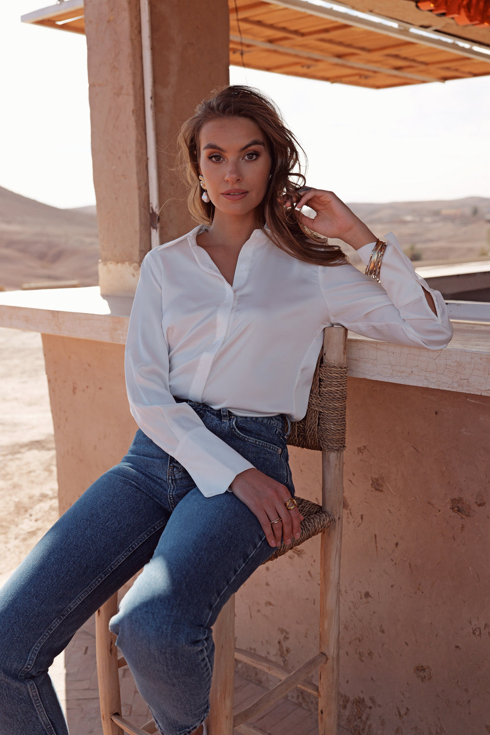 A woman wearing a white long-sleeve button-up shirt and blue jeans sits on a wooden chair in a desert setting with sandy walls and hills in the background.