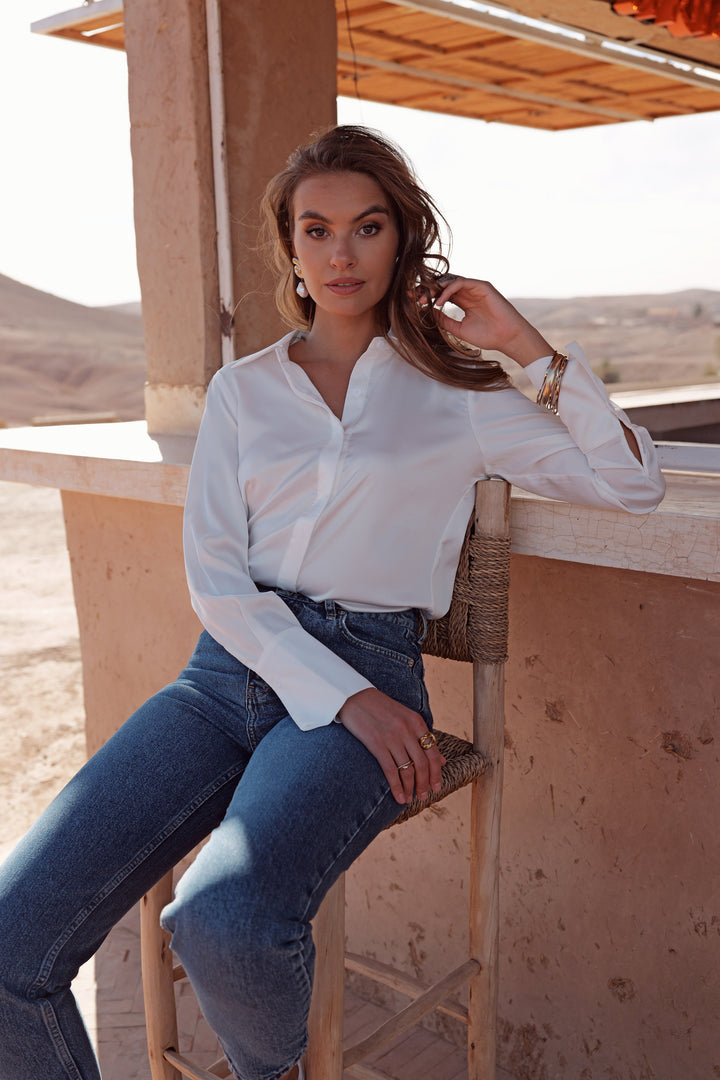 A woman wearing a white long-sleeve button-up shirt and blue jeans sits on a wooden chair in a desert setting with sandy walls and hills in the background.