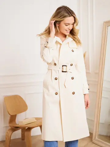 Woman wearing a beige trench coat in a room with a chair and mirror.