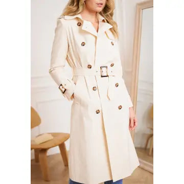 Woman wearing a beige trench coat with gold buttons in an indoor setting.