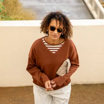 Woman wearing a brown sweater with a striped shirt underneath, sunglasses, and white pants standing outdoors.