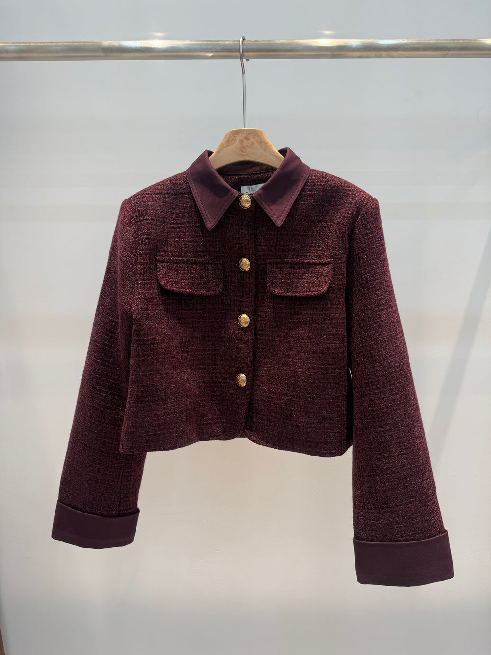A cropped, long-sleeve tweed jacket in a deep burgundy or plum purple color, featuring gold buttons and a matching pointed collar, hanging on a wooden hanger against a plain light gray background.