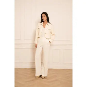 A woman modeling a cream-colored two-piece suit consisting of a cropped jacket and wide-leg trousers, standing against a white paneled wall.