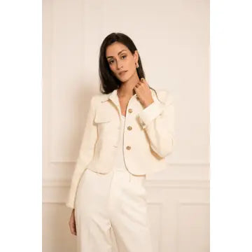 A woman modeling a cropped cream-colored jacket with gold buttons, paired with matching cream trousers, set against a light cream paneled wall.