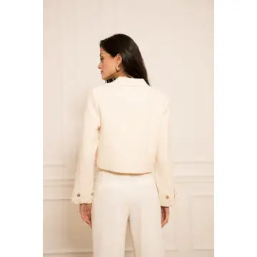 A woman shown from the back wearing a cream-colored cropped blazer and matching trousers against a light beige paneled wall.
