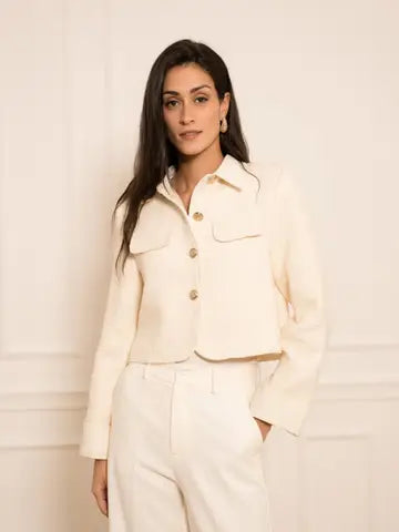 A woman modeling a cropped cream-colored jacket with gold buttons and matching cream trousers, standing against a light cream paneled wall.
