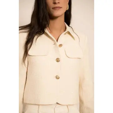 A close-up of a model wearing a cream-colored tweed jacket with gold buttons and matching trousers against a light beige background.