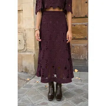 A woman wearing a dark plum lace midi skirt with a matching cropped lace top, standing outdoors in front of a stone wall and wooden door.
