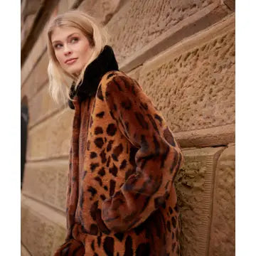 A woman wearing a brown faux fur coat with a black leopard print and a black collar, leaning against a stone wall.