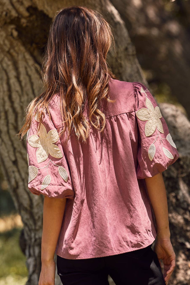 Person wearing a pink blouse with floral embellishments standing against a tree.