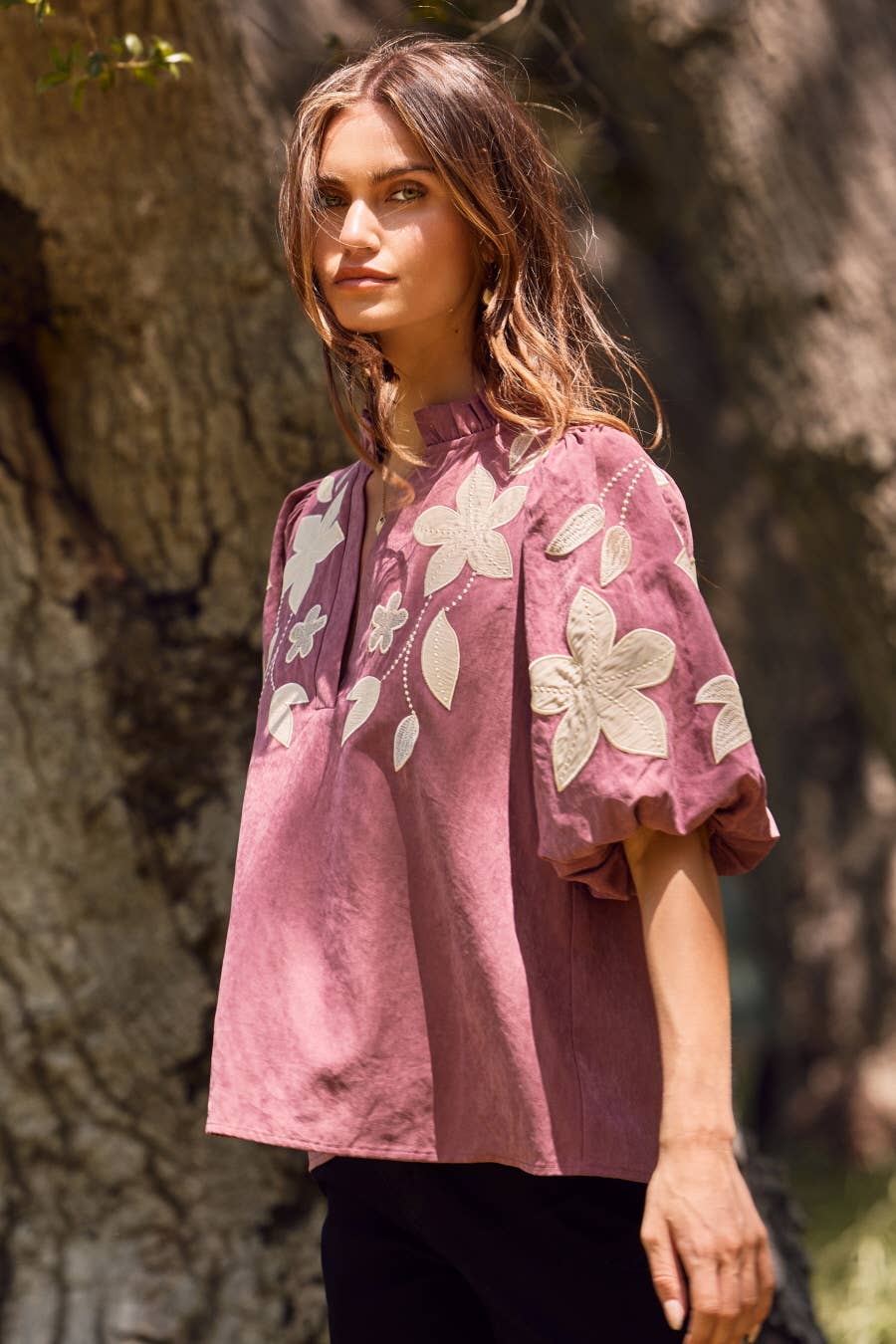 Woman wearing a pink blouse with floral embroidery standing in front of a tree.
