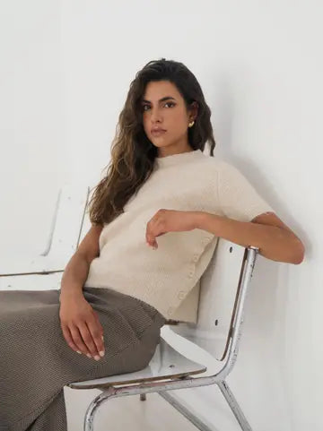 A woman with long wavy brown hair sits on a white chair with a chrome frame, wearing a cream-colored short-sleeve knit top and a textured gray skirt, against a white wall.