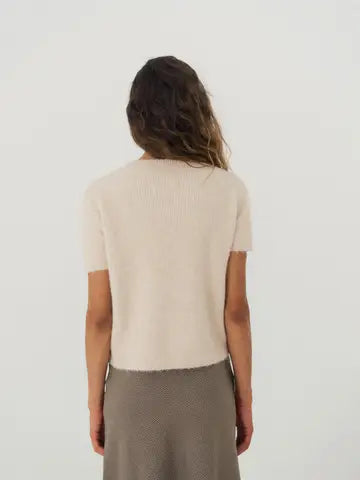 A woman viewed from behind wearing a short-sleeve ribbed knit top in a light cream color and a textured taupe skirt, set against a plain light gray studio background.