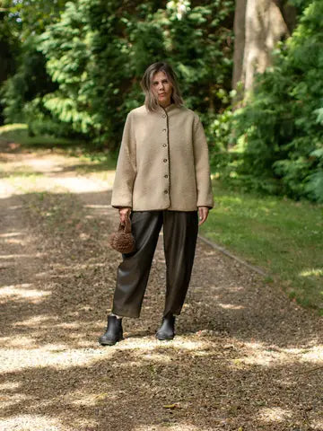 A woman stands on a sun-dappled dirt path in a wooded outdoor setting, wearing a beige cropped button-front jacket with a stand collar, dark brown wide-leg pants, black ankle boots, and holding a small woven brown handbag.