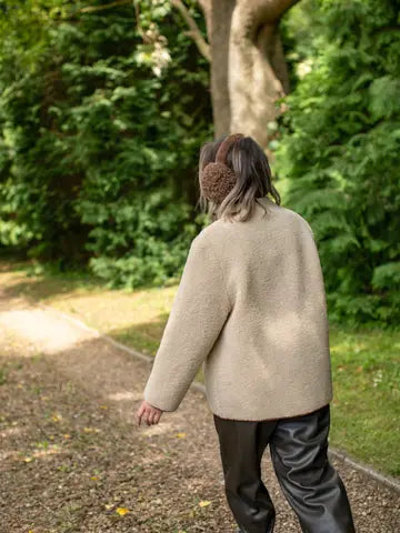 A person viewed from behind wearing a beige sherpa fleece jacket, brown knit earmuffs, and black leather pants, walking on a gravel path in a lush green park.