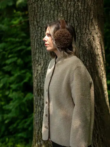 A woman in profile wearing a beige sherpa fleece button-front cardigan and brown fuzzy earmuffs, standing outdoors next to a large tree trunk with green foliage in the background.