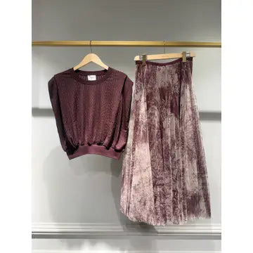 A two-piece women's outfit set hanging on a clothing rack, featuring a textured crewneck sweater in a deep plum purple and a matching pleated midi skirt in a lighter mauve-purple velvet-like fabric.