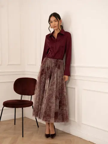 A woman stands in a white paneled room wearing a long-sleeve burgundy satin shirt and a matching dusty mauve velvet tulle midi skirt. She is standing next to a matching burgundy velvet chair.