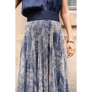 A woman wearing a navy blue sleeveless top with a wide matching waistband and a pleated midi skirt featuring a blue toile-style print on a light beige background.