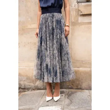 A woman wearing a pleated midi skirt with a blue and gray abstract pattern, paired with a navy top and white pointed-toe heels, standing against a stone wall.