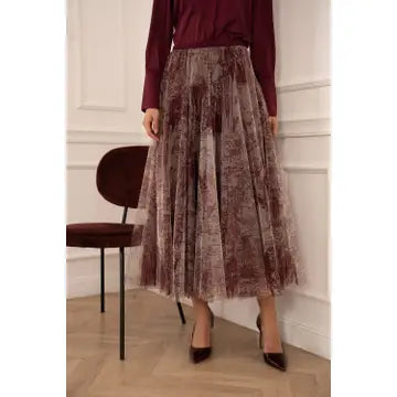 A woman wearing a long-sleeve burgundy top and a pleated tulle midi skirt with a brown and burgundy damask pattern, paired with dark brown pointed-toe heels, standing in a room with a wooden floor and a dark chair.