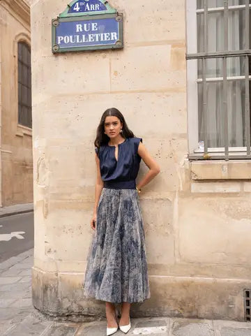 A woman stands outdoors against a beige stone building corner, wearing a sleeveless navy top and a long gray-blue patterned midi skirt. A blue street sign reading Rue Poulletier is visible above her.