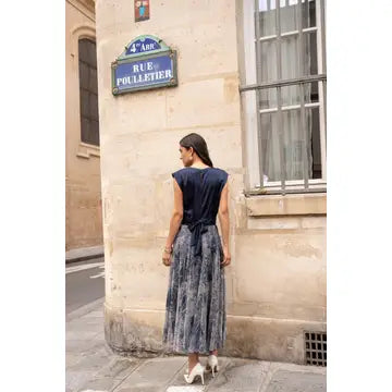 A woman stands on a city sidewalk wearing a sleeveless navy top and a long patterned skirt, photographed from the back against a beige stone building with a blue street sign.
