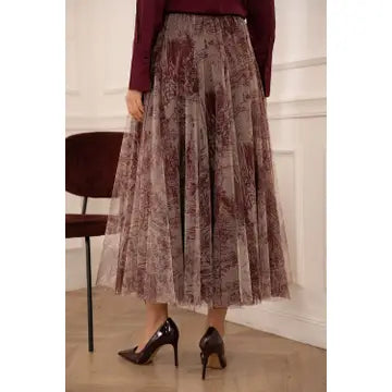 A woman wearing a pleated midi skirt with a brown and mauve floral pattern, paired with a burgundy top and black heels, standing in an indoor setting.