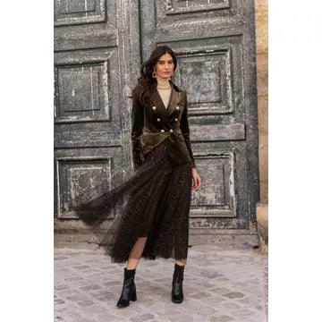 A woman models a dark brown double-breasted jacket and matching tulle midi skirt set with gold buttons, standing in front of a large rustic wooden door.