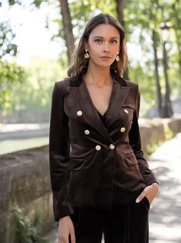A woman wearing a dark brown velvet double-breasted blazer with gold buttons, standing outdoors on a stone pathway with trees in the background.