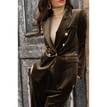 A woman wearing a dark brown velvet double-breasted blazer with gold buttons and matching wide-leg trousers, styled with a beige turtleneck, standing against a rustic gray wooden door.