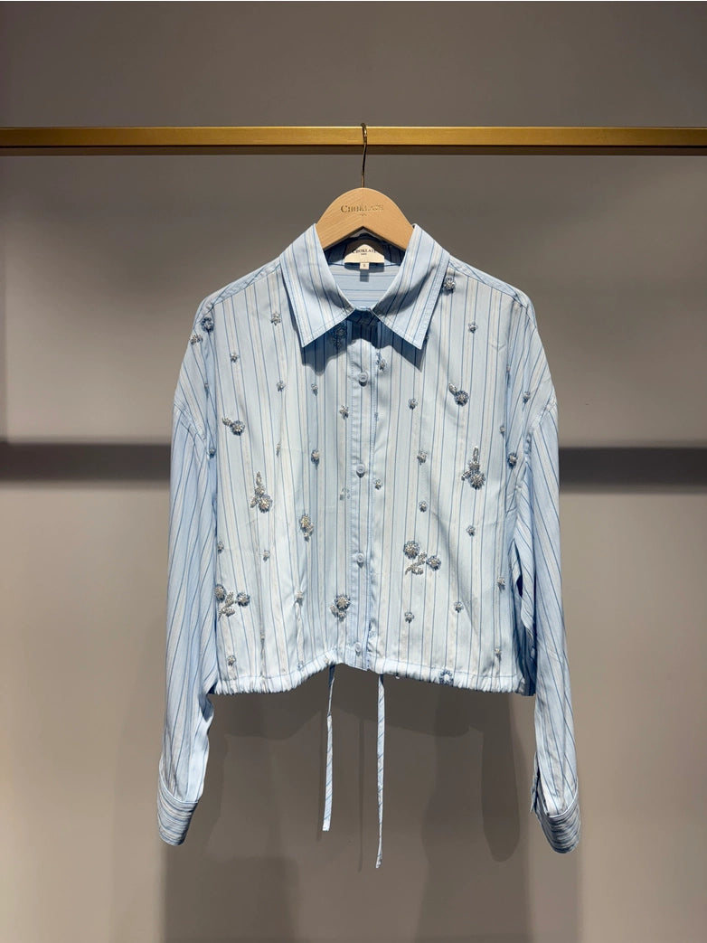 Light blue striped shirt with decorative elements on a hanger against a neutral background