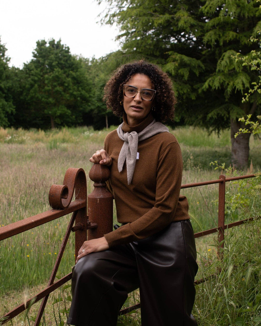Woman sitting on a rusted metal fence in a natural setting with trees and grass.