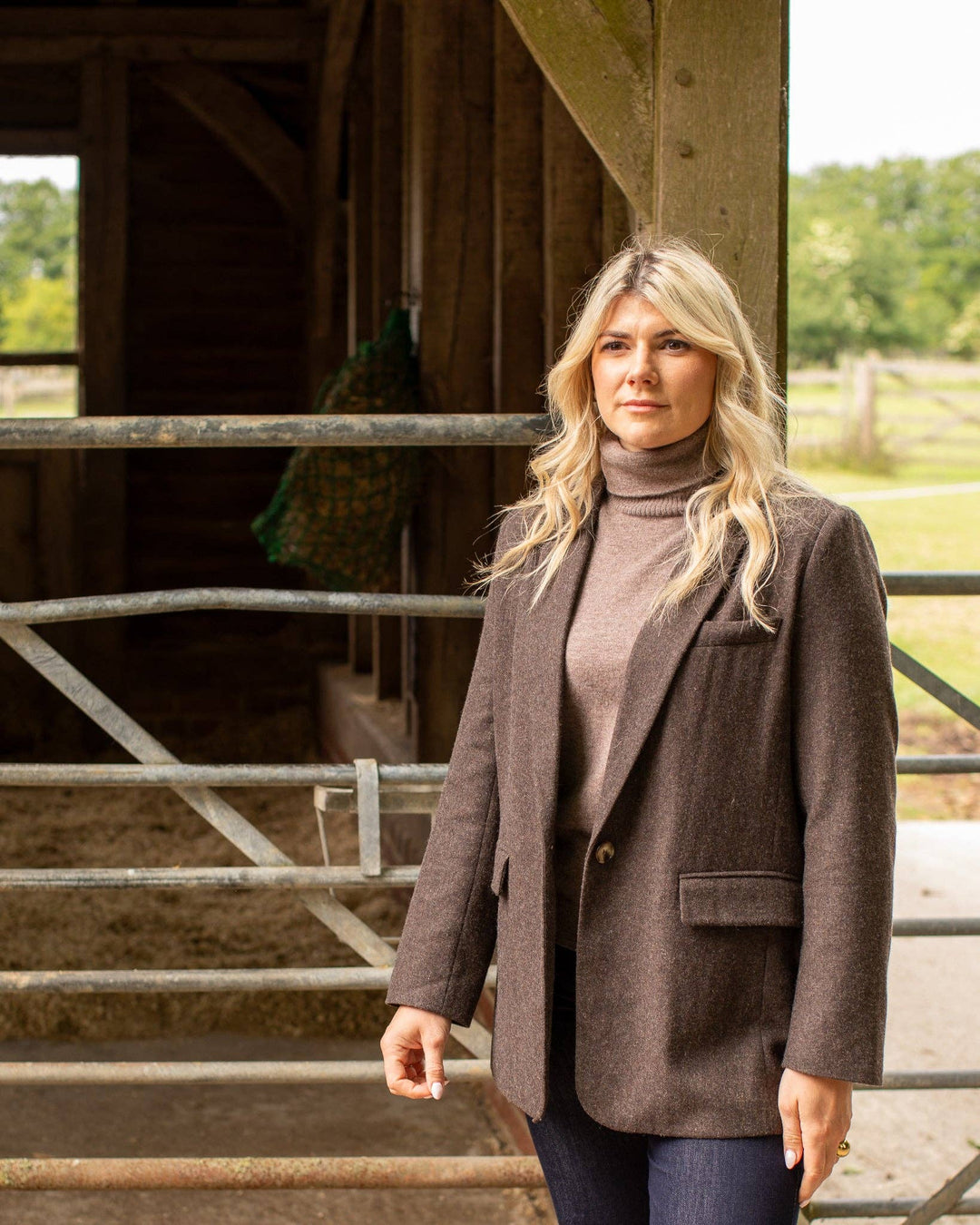 Woman wearing a brown blazer and turtleneck standing in front of a wooden structure.