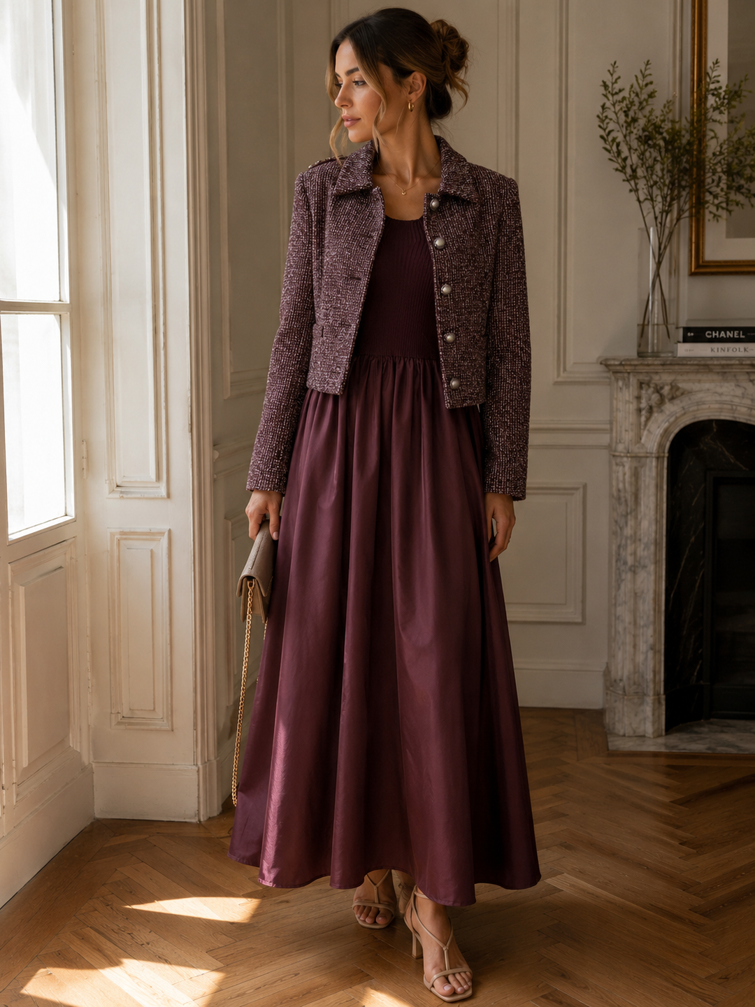 A woman stands in a sunlit interior wearing a long mauve-burgundy dress with a textured cropped jacket, holding a clutch and wearing strappy heels.