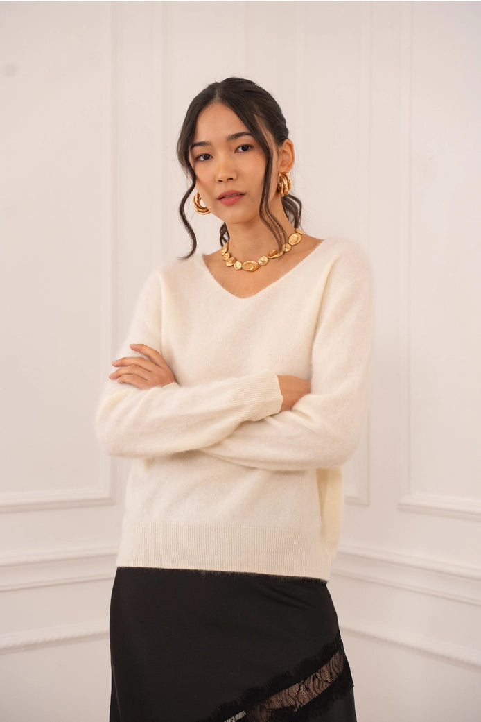 Woman wearing a cream sweater and black skirt with gold jewelry against a white background
