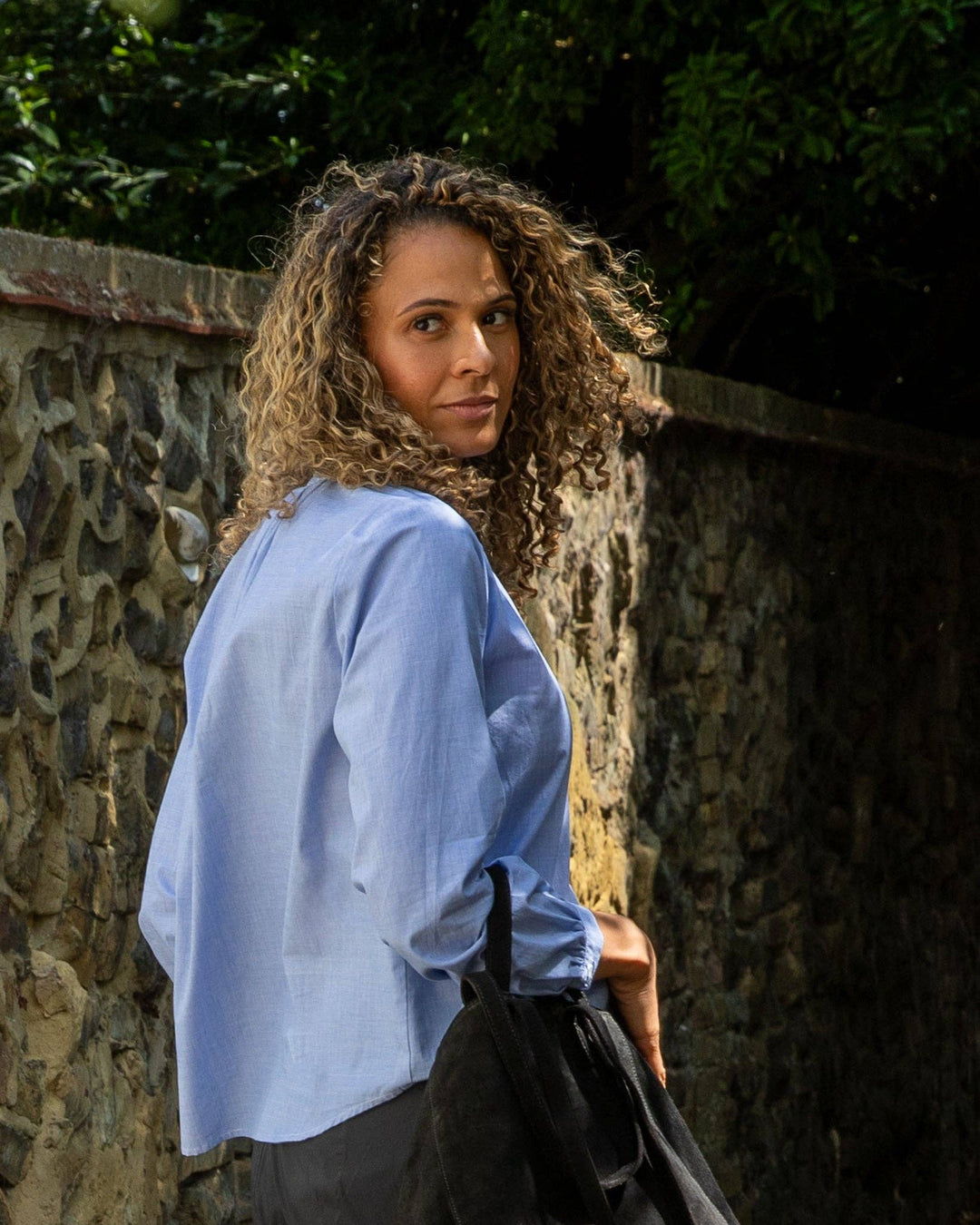 Woman in a light blue shirt standing against a stone wall with greenery.