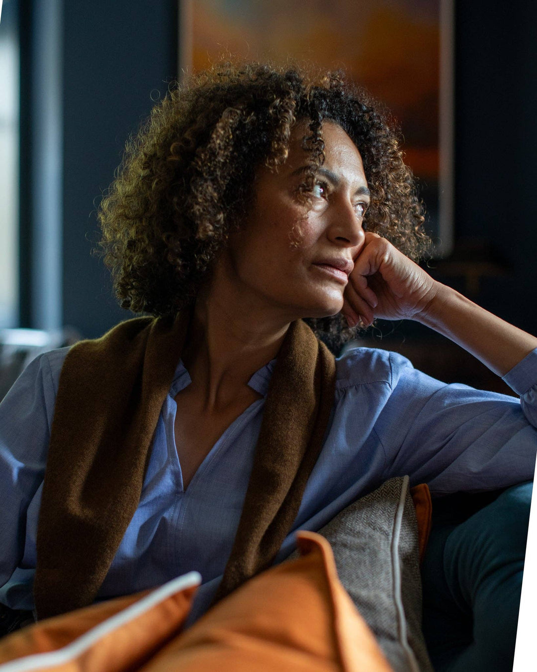 Woman sitting on a couch with a thoughtful expression, wearing a blue shirt and brown jacket.