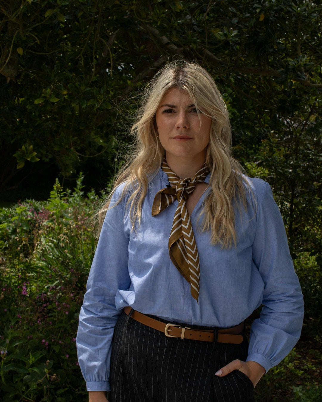 Woman wearing a blue shirt with a brown tie and black pants in a garden setting
