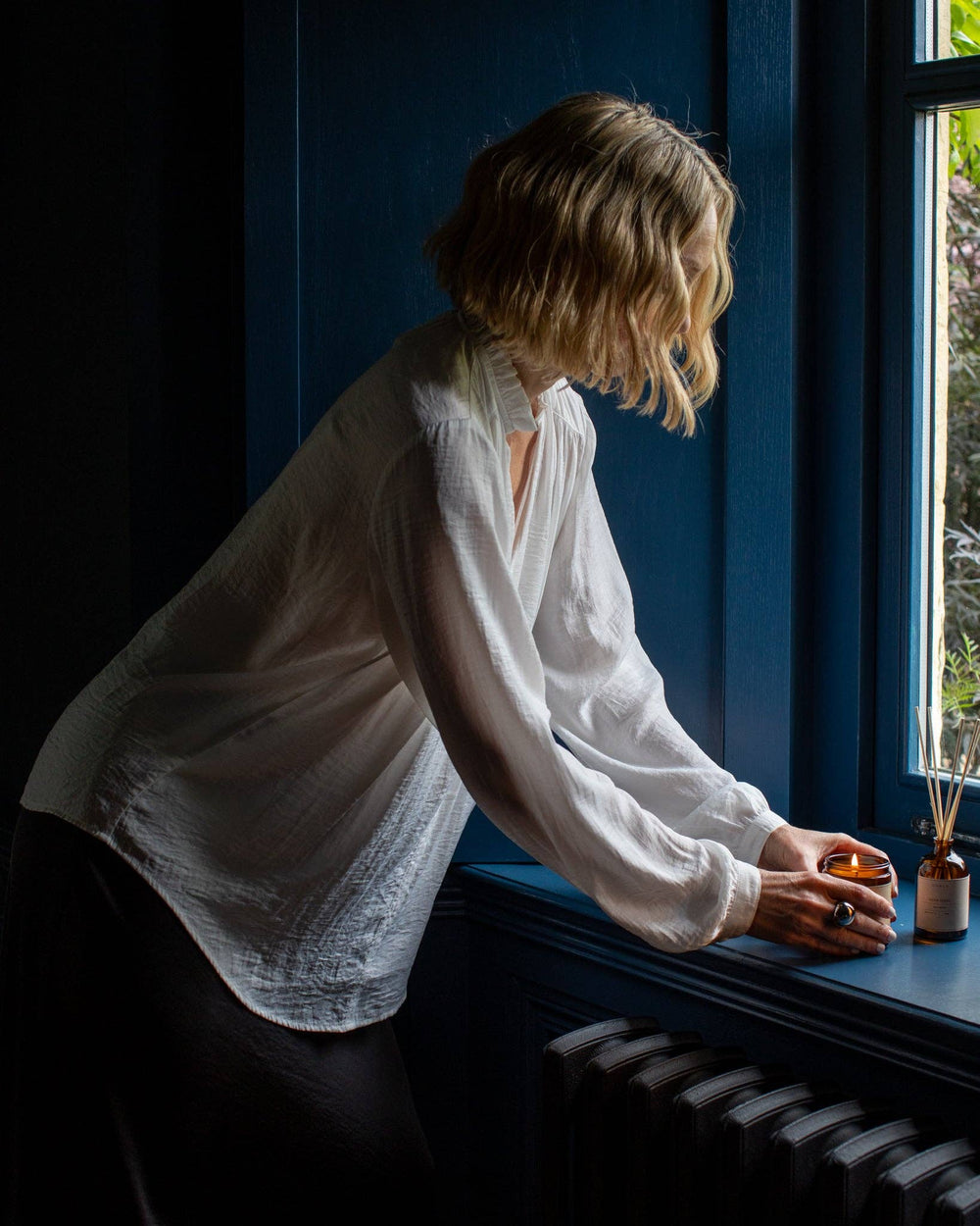 Woman in a white shirt sitting by a window with a candle and small bottle.