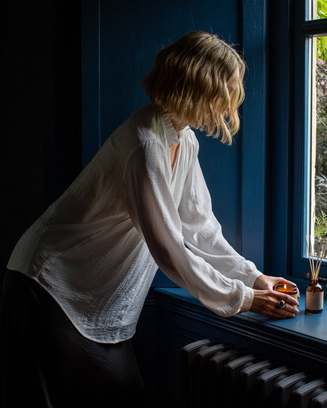 Woman in a white shirt sitting by a window with a candle and small bottle.