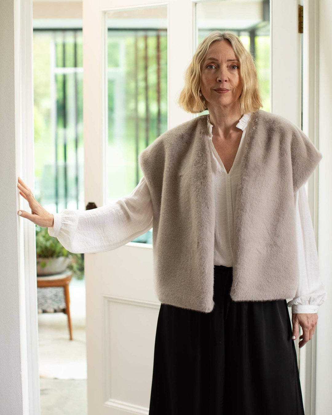 Woman wearing a beige cardigan over a white shirt and black skirt in a bright room.