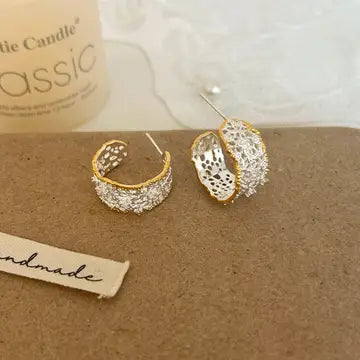 A pair of gold hoop-style earrings with intricate silver filigree and pave crystal detailing, displayed on a brown textured card next to a handmade paper tag.