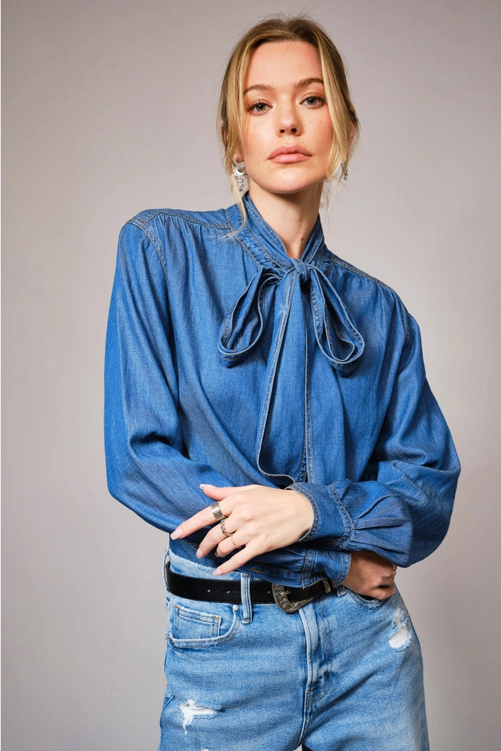 Woman wearing a blue denim blouse with a tie detail and matching jeans against a plain background