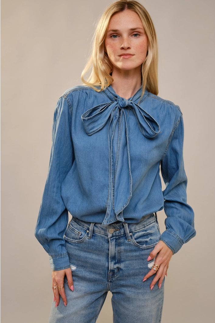 Woman wearing a blue denim blouse with a bow tie and jeans against a beige background