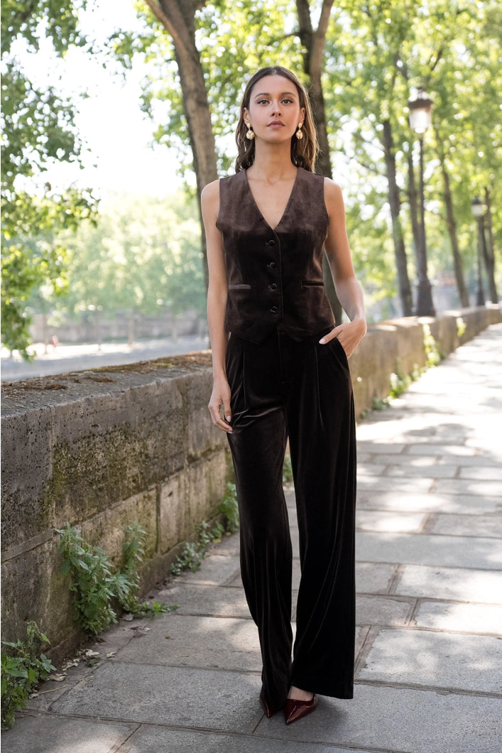 Woman in a brown sleeveless jumpsuit standing on a sidewalk with trees in the background