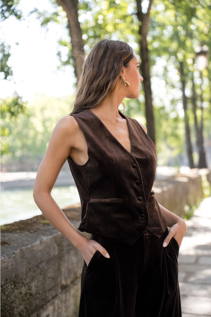 Woman in a brown sleeveless top and black pants standing outdoors with trees in the background
