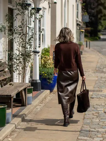 A woman viewed from behind walking down a cobblestone street, wearing a dark brown long-sleeve knit top, a shiny dark brown faux-leather midi skirt, and dark brown knee-high boots, while carrying a dark brown tote bag.
